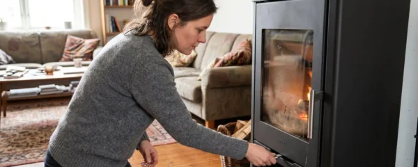Une personne vue de profil ajuste le levier de commande d'arrivée d'air primaire d'un poêle à bois moderne installé dans un salon lumineux